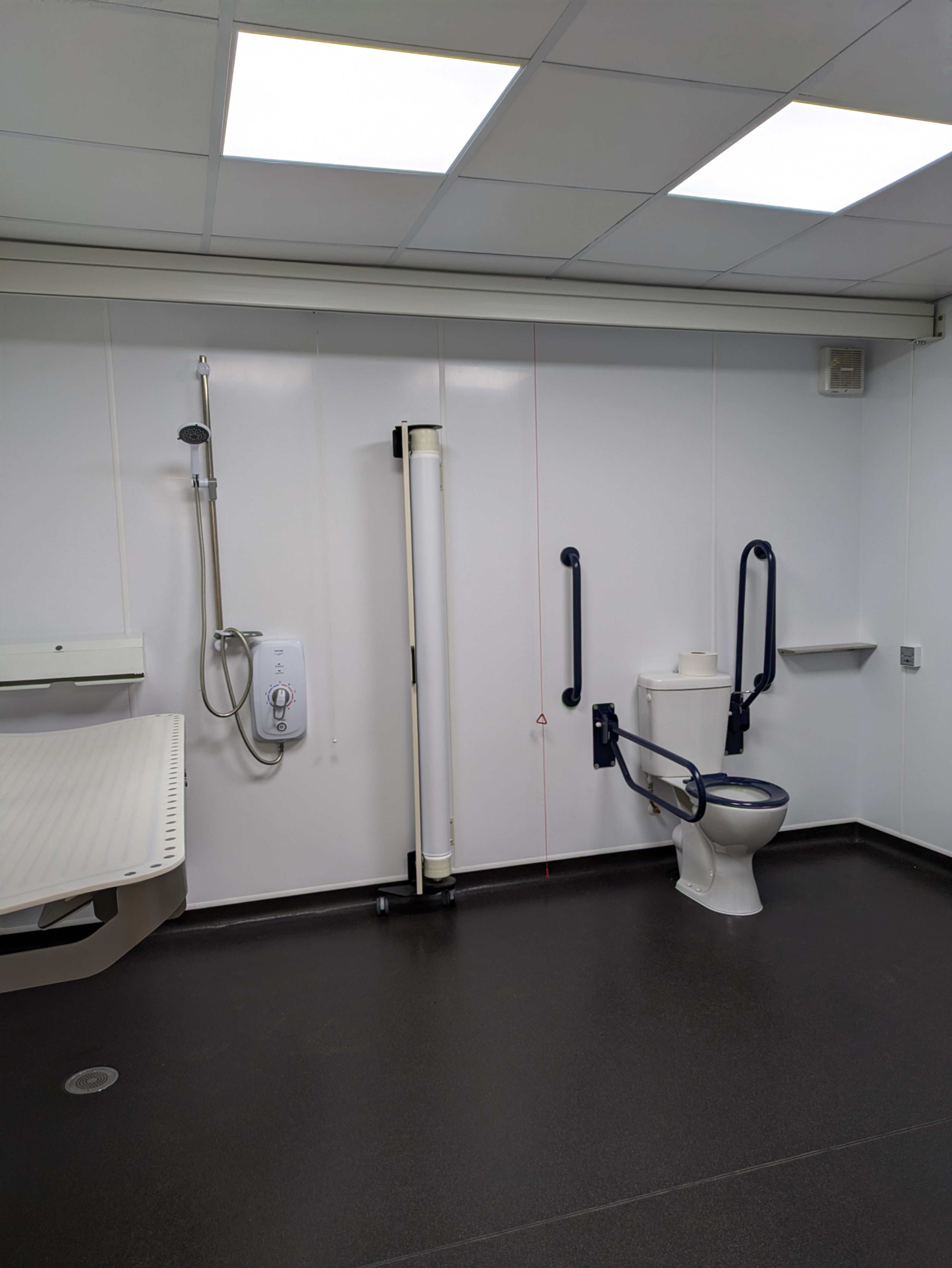 Image shows a changing place facility with black floor and white walls. There is a toilet with handrails, a shower, shower screen and change bed.