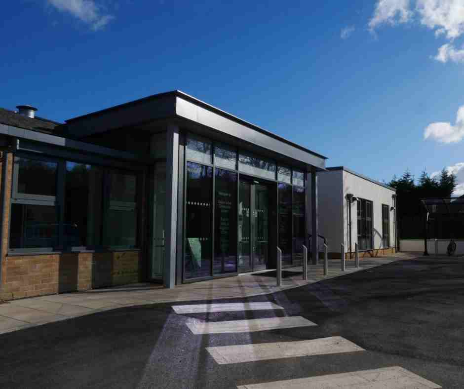 The image shows the front of Oaken Grove Community Centre which is a single storey building with large glass automatic doors at the front. There is a while part of the building to the right of the image and a zebra crossing painted on the tarmac in front.